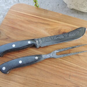 Puede incluir: Un cuchillo y un tenedor con mango negro y hoja con patr&oacute;n. El cuchillo y el tenedor est&aacute;n sobre una tabla de cortar de madera.