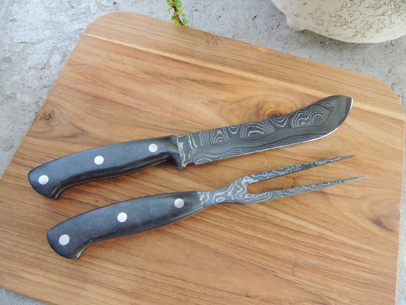 Puede incluir: Un cuchillo y un tenedor con mango negro y hoja con patr&oacute;n. El cuchillo y el tenedor est&aacute;n sobre una tabla de cortar de madera.