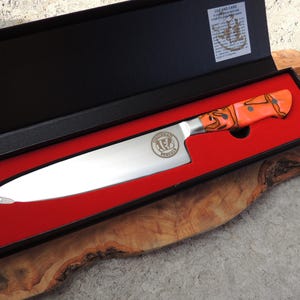 May include: A stainless steel chef's knife with a black and orange handle in a red velvet lined box. The knife has a logo engraved on the blade that says "Fox Valley Knives".