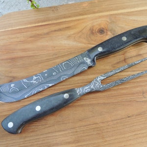 Puede incluir: Un juego de cuchillo y tenedor con mango negro, hoja y dientes con patr&oacute;n. El juego est&aacute; sobre una tabla de cortar de madera.