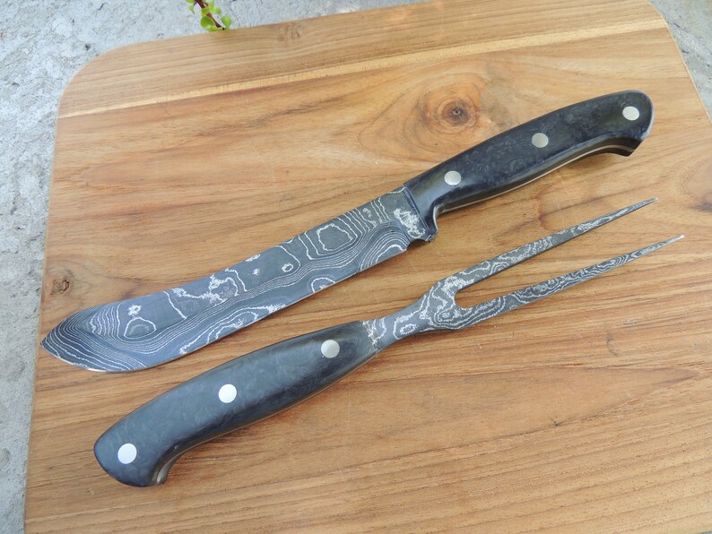 Puede incluir: Un juego de cuchillo y tenedor con mango negro, hoja y dientes con patr&oacute;n. El juego est&aacute; sobre una tabla de cortar de madera.