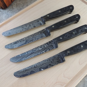 Puede incluir: Cuatro cuchillos de cocina con mango negro y hoja estampada. Las hojas tienen un patr&oacute;n ondulado y veteado en tonos grises.