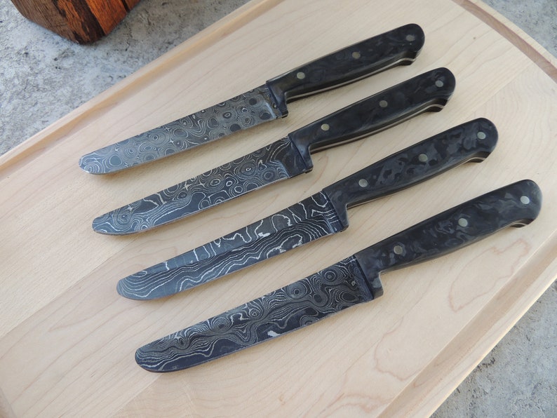Puede incluir: Cuatro cuchillos de cocina con mango negro y hoja estampada. Las hojas tienen un patr&oacute;n ondulado y veteado en tonos grises.