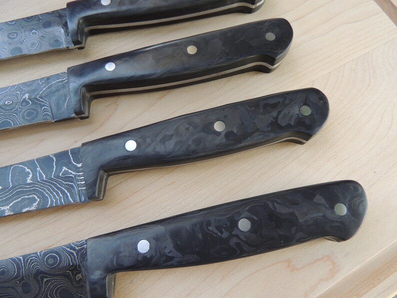 Puede incluir: Cuatro cuchillos de cocina con mangos de fibra de carbono negra y remaches plateados. Las hojas est&aacute;n hechas de acero estampado.