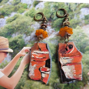 Könnte beinhalten: Handgefertigte Ohrringe mit einem abstrakten Design. Die Ohrringe haben eine rustikale, künstlerische Ästhetik mit orangefarbenen, weißen und schwarzen Details. Sie sind mit bernsteinfarbenen Perlen und einem bronzefarbenen Haken versehen.