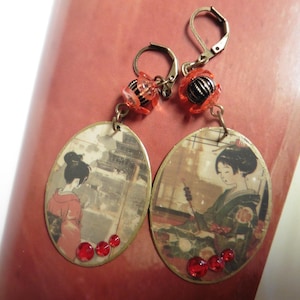 Puede incluir: Pendientes colgantes con colgantes ovalados que representan geishas japonesas. Los pendientes tienen detalles en rojo, incluyendo pequeñas cuentas y gemas rojas. El metal tiene un tono dorado. Cierre de palanca.