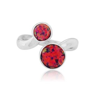 Op de afbeelding: Een zilveren ring met twee ronde, bezel-set, rode opaalstenen. De opalen hebben een levendige mix van rode, paarse en groene tinten. De band van de ring heeft een uniek, vloeiend ontwerp.