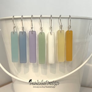 May include: A collection of rectangular earrings in various pastel colors, including blue, purple, green, white, yellow, and orange. The earrings are suspended from silver-colored hooks and displayed against a white bucket.