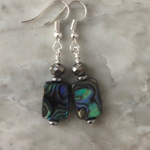 Può includere: Orecchini argentati con pendenti rettangolari in conchiglia di abalone. I pendenti iridescenti mostrano sfumature di blu, verde e nero. Ogni orecchino presenta una perla sfaccettata e una piccola perla d'argento.