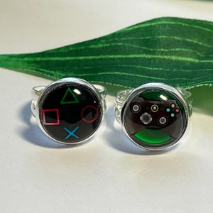 May include: Two silver rings with black and green designs. One ring features the PlayStation controller buttons, a square, circle, triangle, and X. The other ring features a green Xbox controller with black buttons.