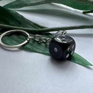 May include: A keyring featuring a glossy black six-sided die with white pips. The die is attached to a silver-toned metal split ring by a short chain. The die shows the numbers one, three, and four clearly on its visible faces, with a smooth finish.