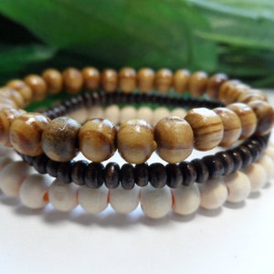 May include: Three stacked bracelets made of wooden beads. The top bracelet is made of brown and beige striped beads. The middle bracelet is made of dark brown beads. The bottom bracelet is made of light brown beads.