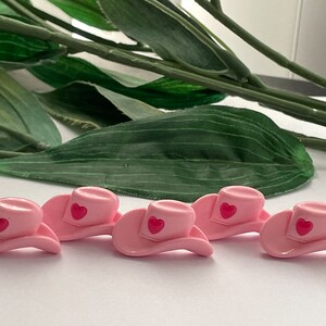 May include: Six pink cowboy hats with a red heart design. The hats are arranged in a row on a white surface.