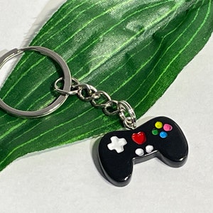 May include: Black video game controller keychain with colorful buttons and a silver key ring. The controller has a white cross and a red heart. The keychain is on a green leaf.
