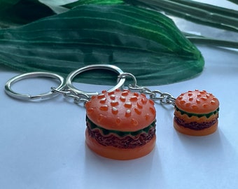 May include: Two burger-shaped keychains. The burgers have orange buns with white sesame seeds, green lettuce, and brown patties. The keychains are attached to silver key rings and chains. The background is a light surface with a green leaf.