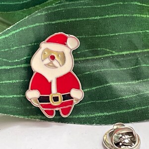 May include: A silver-toned enamel pin featuring a cartoon Santa Claus wearing a red suit with a white trim and a black belt. The Santa Claus has a white beard and a red nose.
