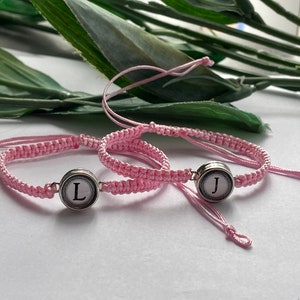 May include: Two pink friendship bracelets with silver charms featuring the letters "J" and "L".