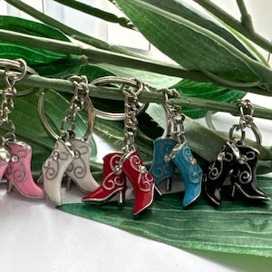 Könnte beinhalten: Sechs bunte schlüsselanhänger in Form von Stiefeln mit silbernen Schlüsselringen. Die Stiefel sind in Pink, Weiß, Rot, Blau und Schwarz mit silbernen Akzenten.