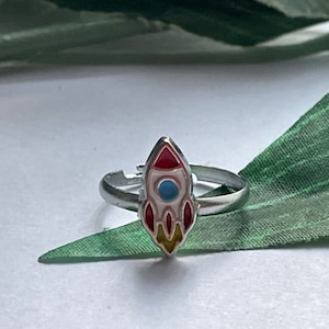 May include: A silver-toned ring featuring a colorful rocket ship design. The rocket has red, white, blue, and yellow enamel details. The ring is adjustable and sits on a green leaf.