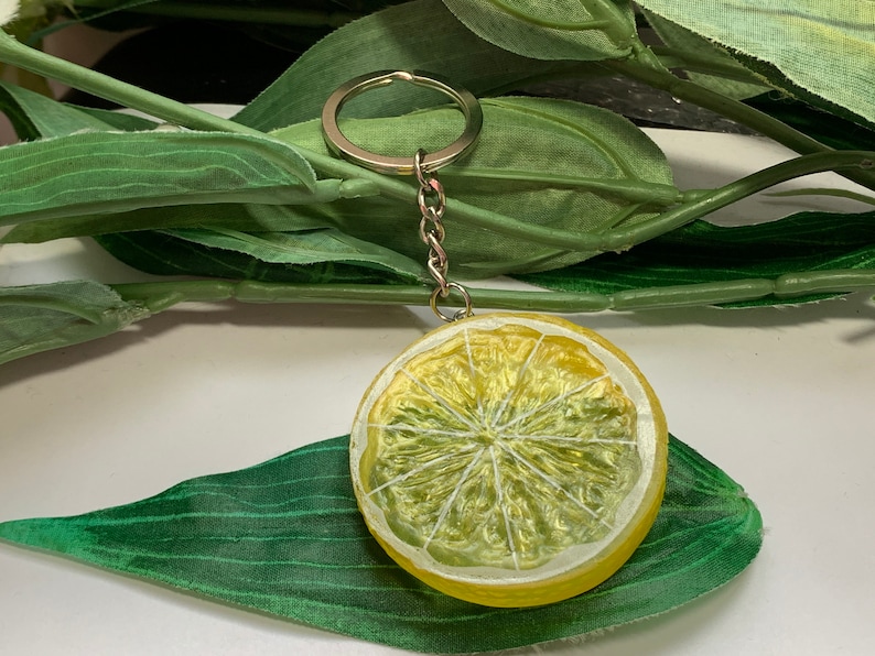May include: A yellow lemon slice keychain with a silver chain and keyring. The lemon slice has a white rind and detailed segments. The keychain is set against a backdrop of green leaves.
