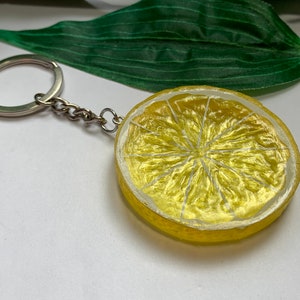 May include: A lemon slice keychain with a silver chain and ring. The lemon slice is yellow with white details, resembling a real lemon. The keychain is a fun accessory.