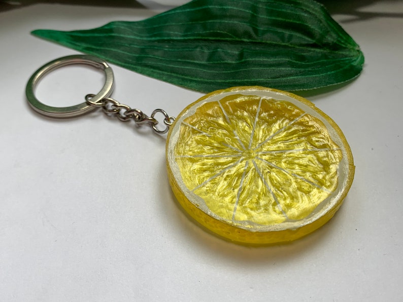 May include: A lemon slice keychain with a silver chain and ring. The lemon slice is yellow with white details, resembling a real lemon. The keychain is a fun accessory.