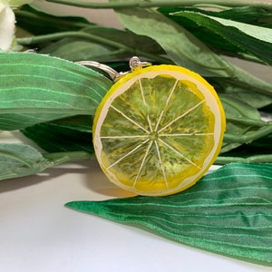 May include: A yellow and green lemon slice keychain. The keychain is round with a painted design of a lemon slice, featuring white segments and a yellow rind. The background includes green leaves.