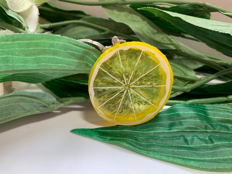 May include: A yellow and green lemon slice keychain. The keychain is round with a painted design of a lemon slice, featuring white segments and a yellow rind. The background includes green leaves.