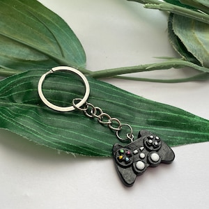 May include: Black and white video game controller keychain with a silver key ring and chain. The keychain is on a green leaf.