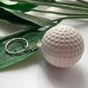 May include: A white golf ball keyring with a silver ring and chain. The golf ball has a dimpled surface and is attached to the keyring with a small plastic piece. The background includes green leaves.