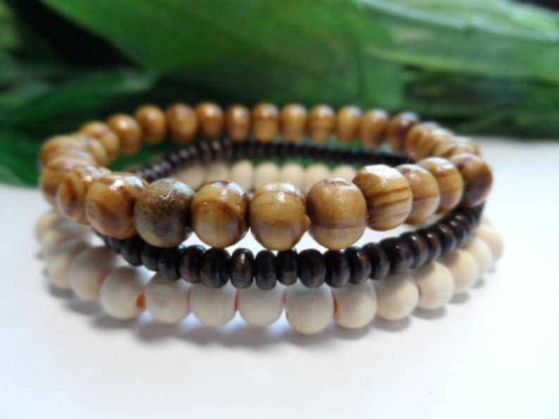 May include: Three stacked bracelets made of wooden beads. The top bracelet is made of brown and beige striped beads. The middle bracelet is made of dark brown beads. The bottom bracelet is made of light brown beads.