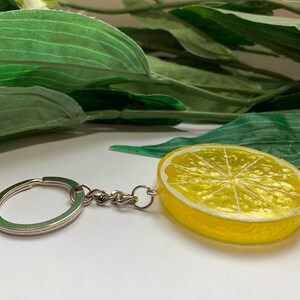 May include: A lemon slice keychain with a silver-colored keyring and chain. The lemon slice is yellow with a white rind and a detailed interior. The background includes green leaves.