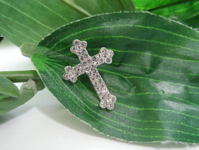 May include: A silver cross with clear rhinestones on a green leaf background.