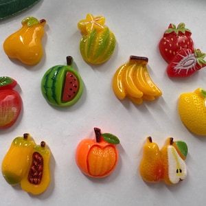 May include: A selection of fruit-shaped decorations in various colours. The collection includes a red apple, a green and red watermelon slice, bananas, strawberries, a lemon, and other fruits. These items are likely used for decoration.
