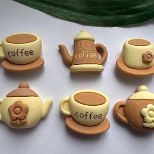 May include: A collection of miniature coffee-themed decorations. The set includes coffee cups, saucers, and teapots in shades of brown and cream. Some pieces feature the word "coffee" and floral accents. These decorative items are likely for crafts or embellishments.