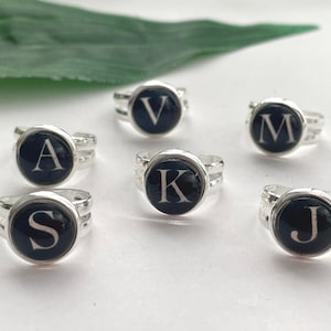 Op de afbeelding: Zilverkleurige ringen met zwarte cirkelvormige vlakken, elk met een andere witte letter. De zichtbare letters zijn A, J, K, M, S en V. De ringen liggen op een wit oppervlak, met een groen blad op de achtergrond.