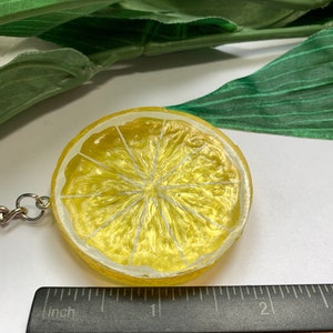 May include: A yellow lemon slice keychain with a silver chain. The keychain is round with a painted design of a lemon slice. The lemon slice has a yellow rind and a white interior with white lines. The keychain is next to a ruler.