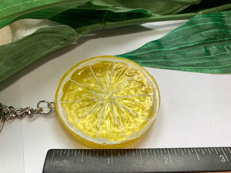 May include: A yellow lemon slice keychain with a silver chain. The keychain is round with a painted design of a lemon slice. The lemon slice has a yellow rind and a white interior with white lines. The keychain is next to a ruler.