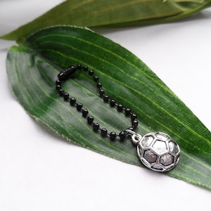 May include: A silver football charm with a black ball chain. The football has a detailed, textured surface. The charm is displayed on a green leaf against a white background.