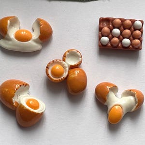 May include: A collection of five miniature resin egg-themed charms. The set includes four individual pieces showing cracked brown shells with yellow yolks and white egg whites, and one rectangular brown carton containing fifteen small eggs.