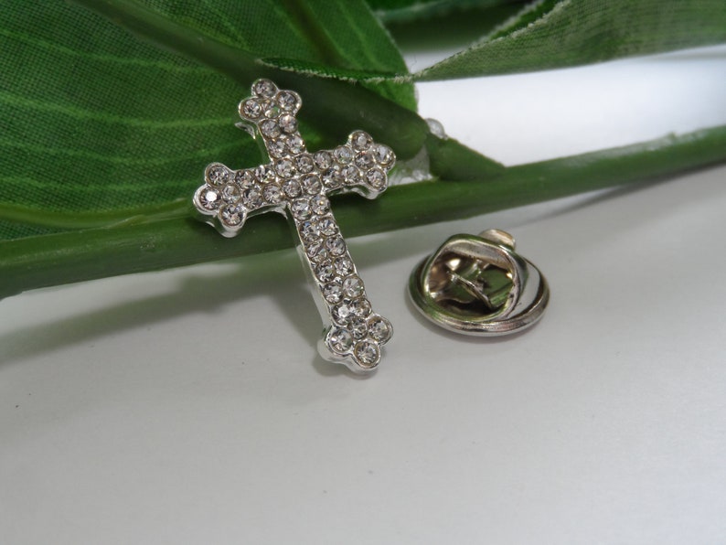May include: A silver cross pin with clear rhinestones. The pin has a safety clasp on the back.