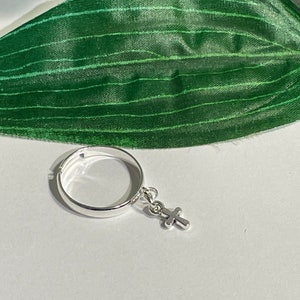 May include: A silver ring with a small cross charm. The ring is simple and shiny, with a small cross pendant hanging from the band. The background is a white surface with a green leaf.