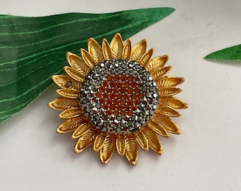 Könnte beinhalten: Eine goldfarbene Sonnenblumenbrosche mit einem Zentrum aus schwarzen und orangefarbenen Strasssteinen.