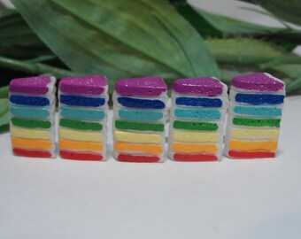 May include: Five miniature rainbow-colored cake slices. Each slice features layers of red, orange, yellow, green, blue, and purple. The top layer is a textured purple, and the sides are white. The cakes are arranged on a white surface.