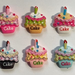 Può includere: Sei ciondoli colorati a forma di cupcake con candele. Ogni cupcake ha una base, una glassa e dettagli decorativi di colore diverso. La parola "Cake" è stampata sulla base di ogni cupcake. Questi ciondoli sono probabilmente per l'artigianato o la creazione di gioielli.