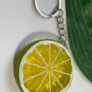 May include: A lime slice keychain with a silver keyring and chain. The lime slice is painted with a bright yellow center and a green rind, outlined in white. The keychain is a fun accessory.