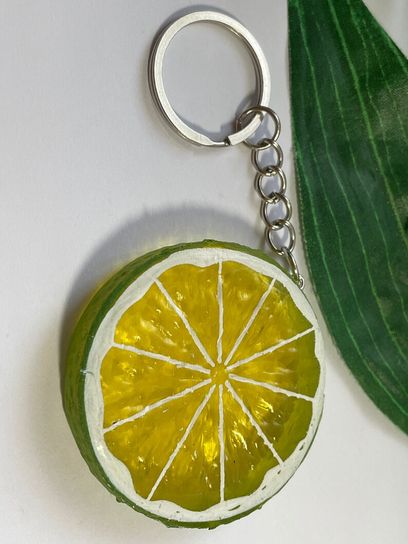 May include: A lime slice keychain with a silver keyring and chain. The lime slice is painted with a bright yellow center and a green rind, outlined in white. The keychain is a fun accessory.