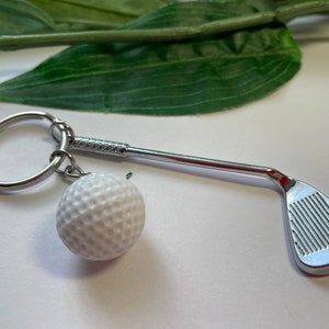 Può includere: Un portachiavi a forma di mazza da golf argentata con un ciondolo a forma di palla da golf bianca. Il portachiavi ha un anello argentato per attaccarlo alle chiavi.