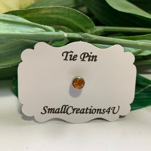 Op de afbeelding: Een stropdaspin met een kleine, ronde, amberkleurige steen. De pin is op een witte kaart met de tekst "Tie Pin" en "SmallCreations4U".