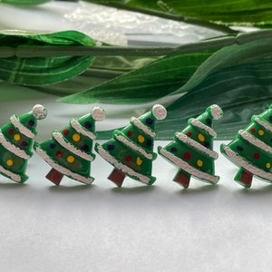 May include: Five green Christmas tree ornaments with white trim, colorful ornaments, and a white ball on top. The trees have brown bases and are arranged in a row. The background includes green leaves.